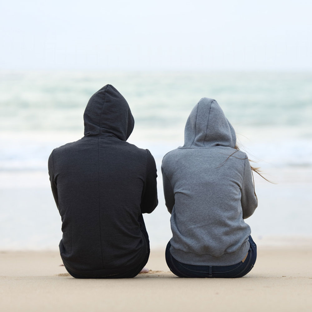 Two people in hoodies sitting on the beach, discussing signs of depression - Uncover Counseling.
